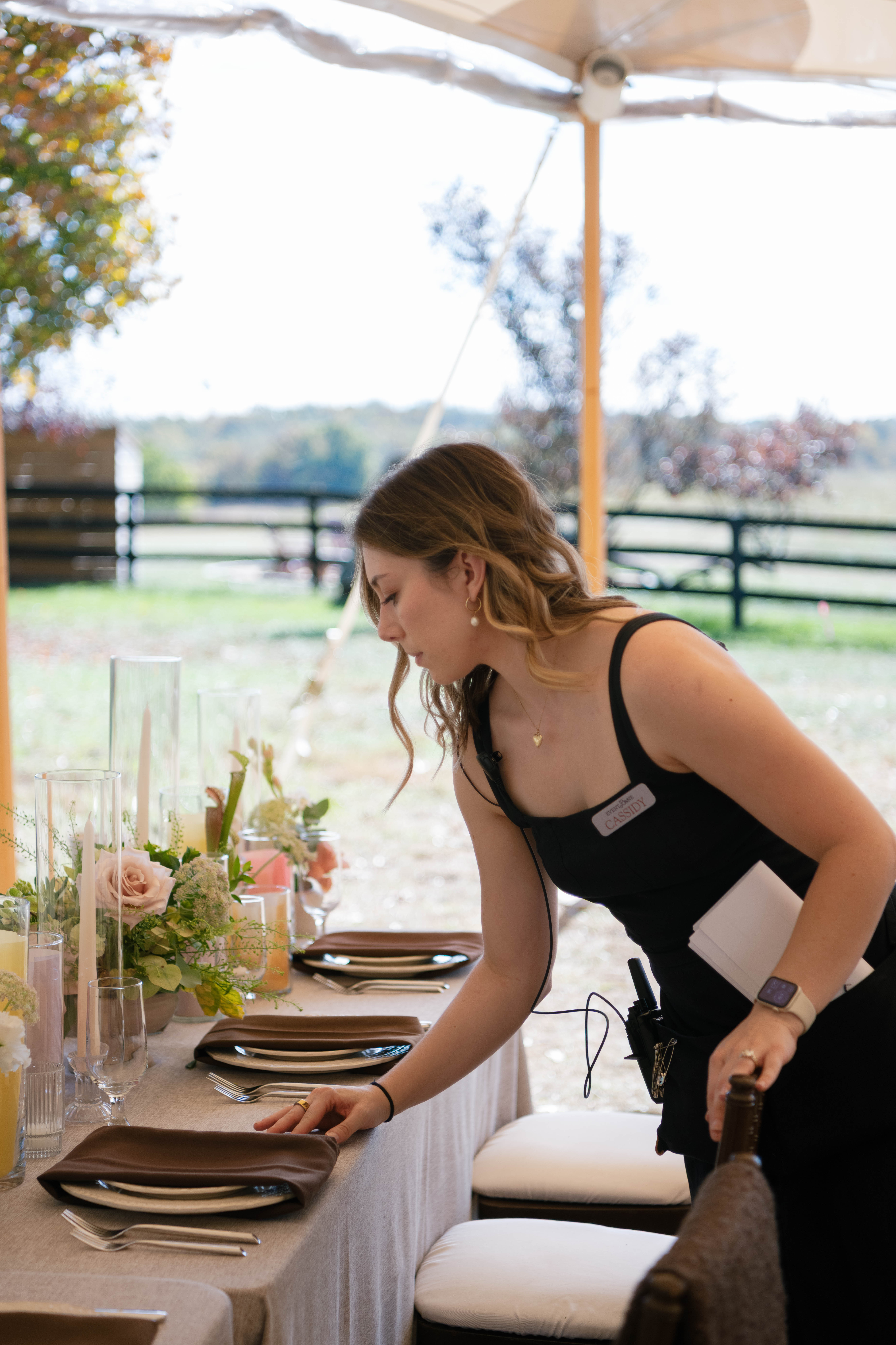Woman adjusts a place setting at a brown and neutral set wedding table in an outdoor tent